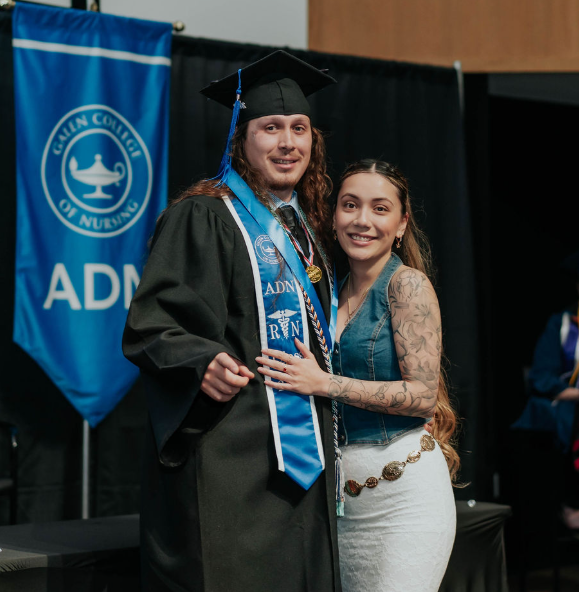 Indoors at a graduation ceremony, a graduate in a cap and gown stands beside another person while posing for a photo. A blue banner in the background reads “Galen College of Nursing ADN RN,” and seated individuals in academic regalia are visible applauding.