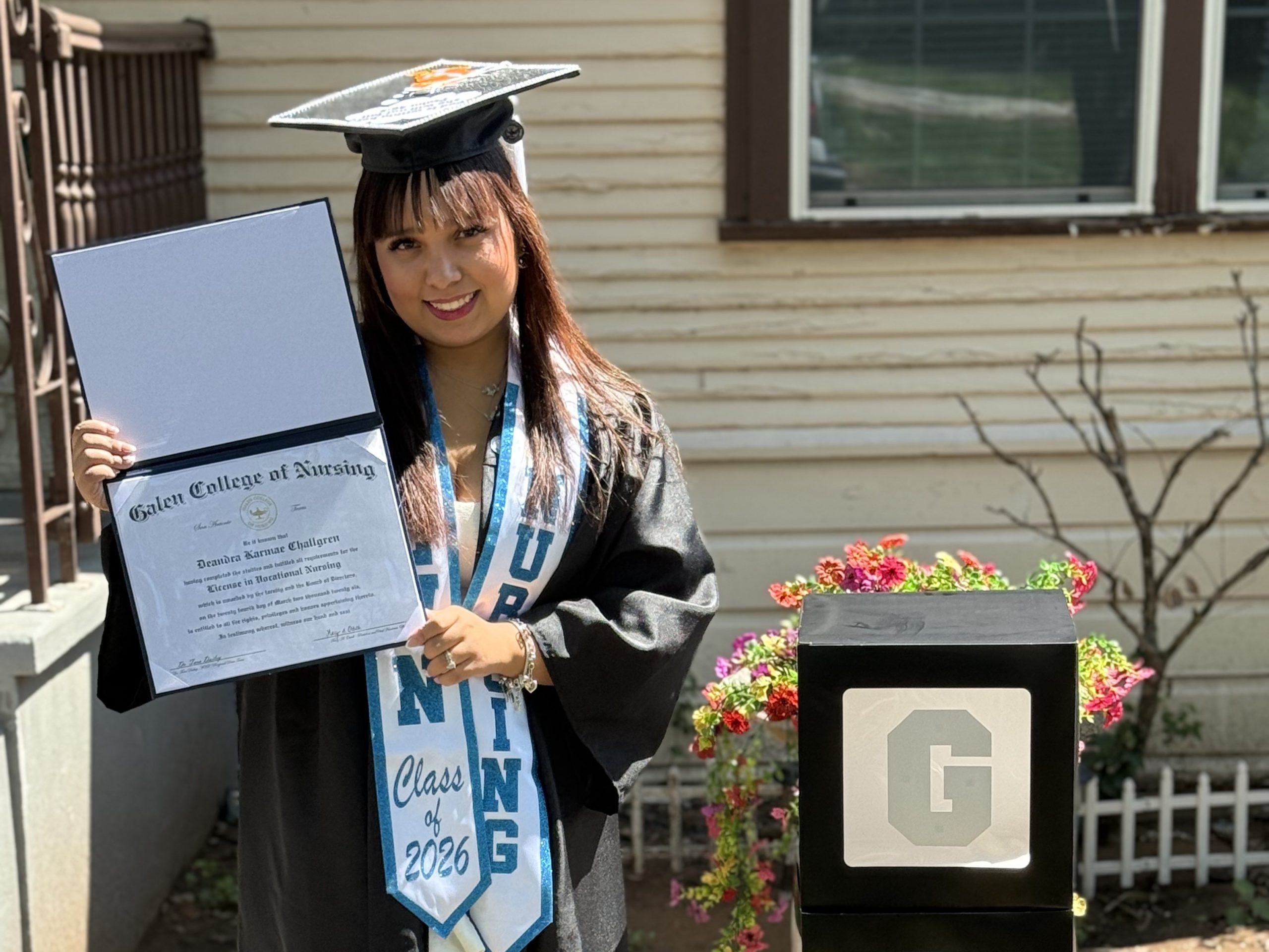 Person wearing a graduation cap and gown holds an open diploma from Galen College of Nursing while standing outdoors near a house and flower bed.