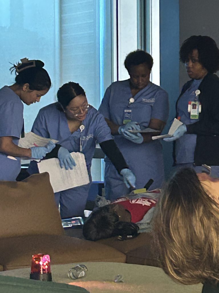 Nursing students in blue scrubs work together during a disaster simulation, reviewing notes while providing care to a patient mannequin on a stretcher