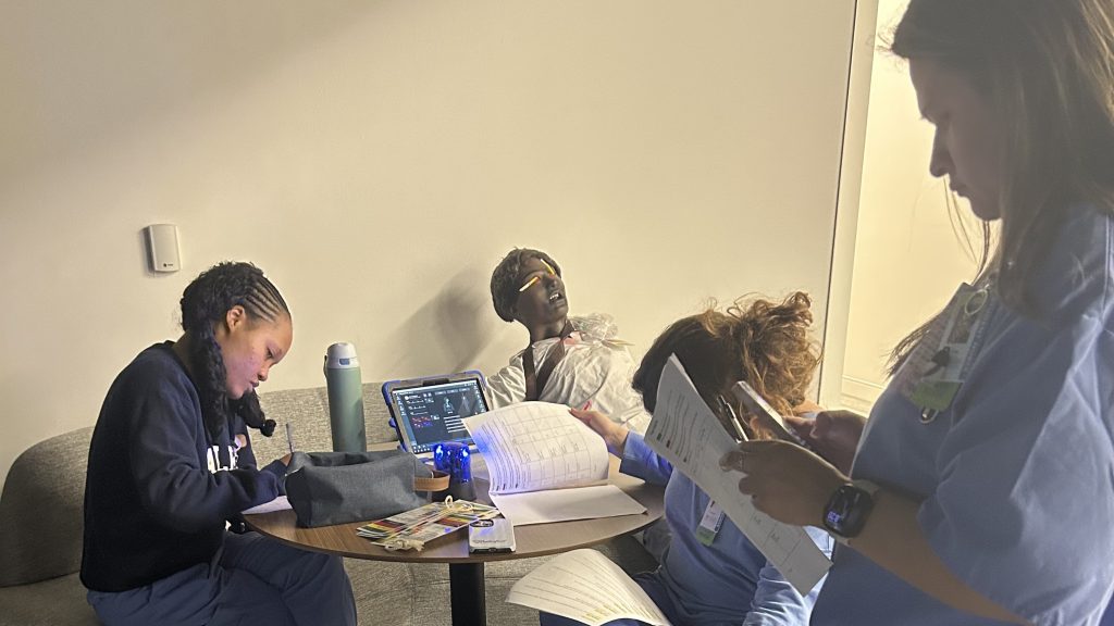 Nursing students review patient charts and take notes at a table beside a mannequin used for simulation training in a clinical learning environment.
