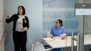 An instructor writing on a white board in a study room, while a student in scrubs watches her