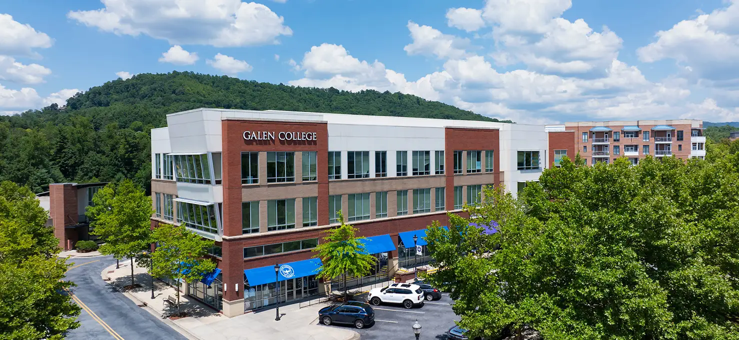 An aerial view of the modern Galen College building in Asheville, North Carolina, nestled among green trees with mountains in the background.
