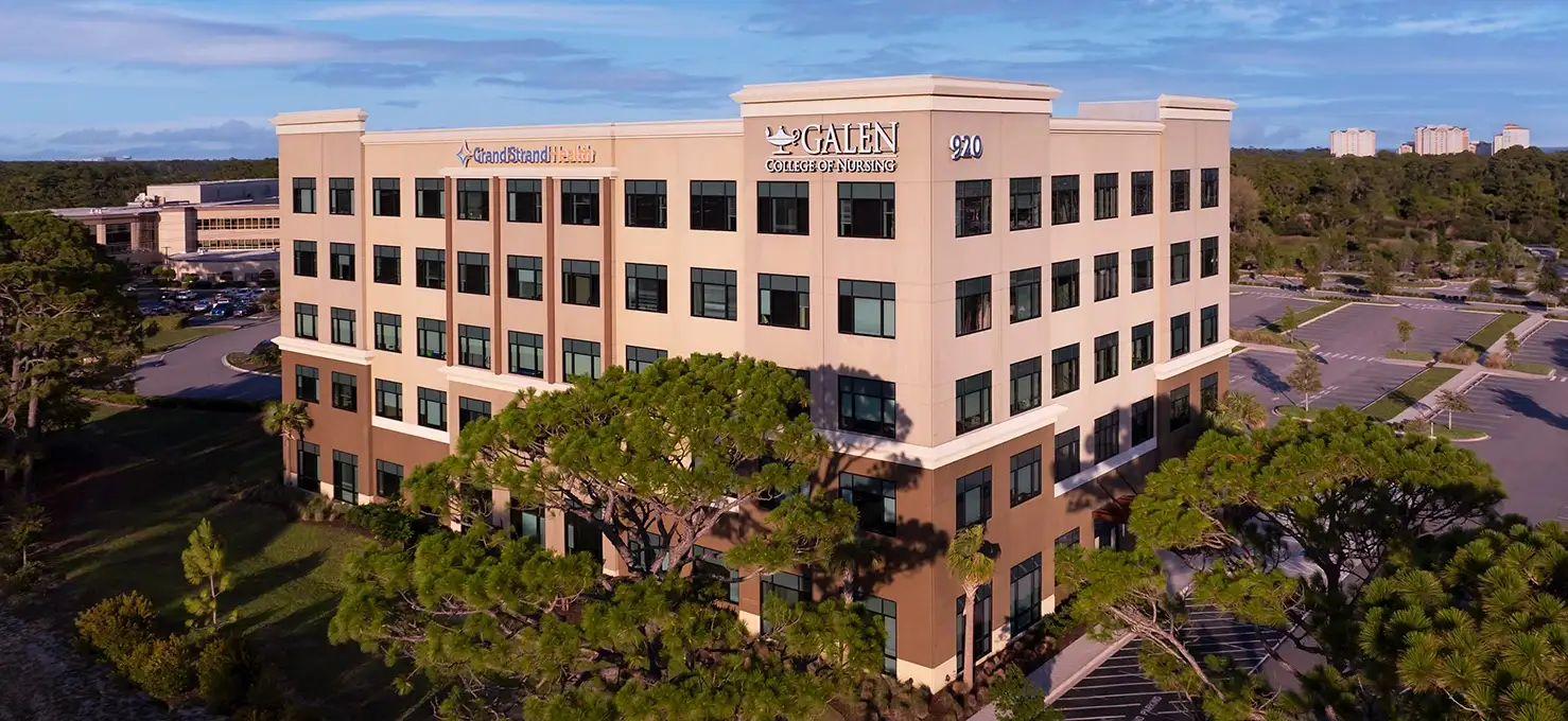 An aerial view of a multi-story Galen College of Nursing building with a tan and brown facade, surrounded by trees and a parking lot, under a partly cloudy sky.