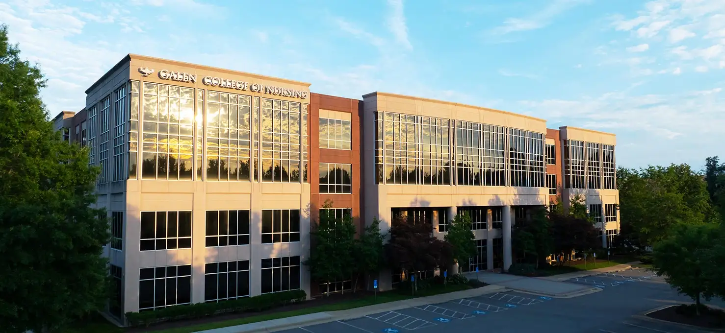 An exterior view of a modern, multi-story building labeled 'GALEN COLLEGE OF NURSING' on its facade. The building has large windows reflecting the golden sunlight and is surrounded by green trees and a paved parking lot under a blue sky.