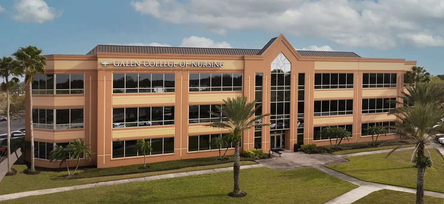 An exterior view of the 'Galen College of Nursing' building in Sarasota, FL, a large, multi-story structure with tan and yellow-ochre colored facades and numerous reflective windows. Palm trees and green lawns surround the building, and a partly cloudy sky is visible overhead.