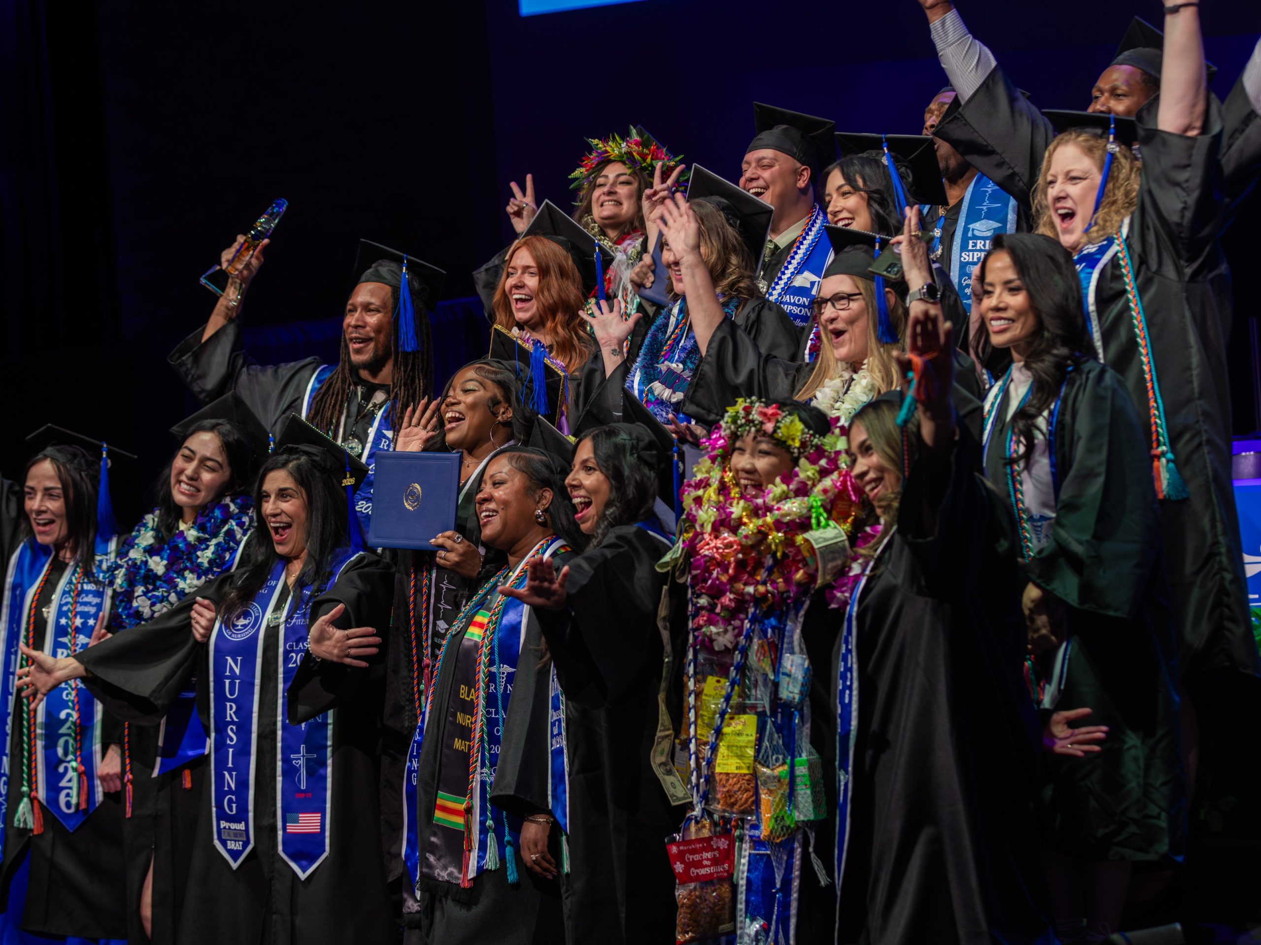 A large group of graduates in caps and gowns stand together on stage during a commencement ceremony, many raising their hands and holding diplomas, wearing honor cords and nursing stoles, with stage lighting and a dark backdrop behind them.