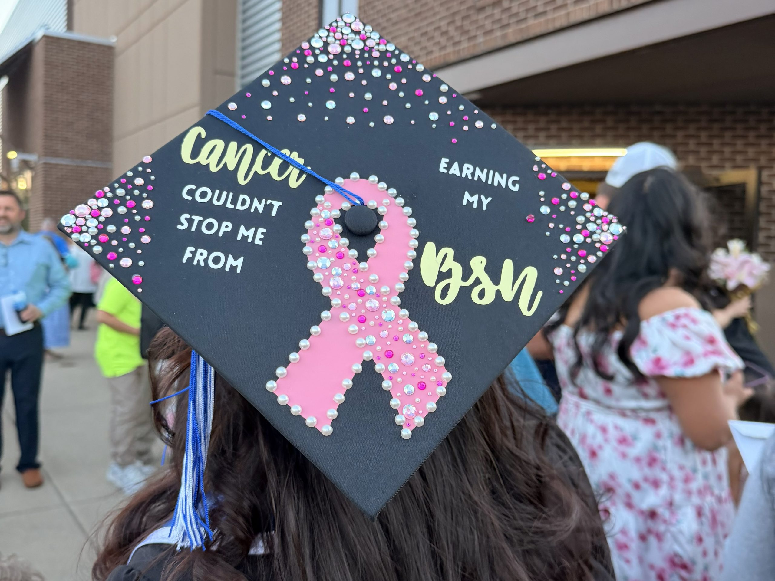 Close-up of a decorated graduation cap featuring a pink ribbon made of rhinestones and pearls, with text that reads “Cancer couldn’t stop me from earning my BSN.” The cap is worn outdoors at a graduation ceremony, with people and a building blurred in the background.