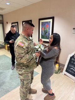 Two people stand facing each other in a hospital hallway or room, holding a bouquet, while another person stands nearby. The setting includes framed artwork on the wall and a candle, suggesting a small ceremony or gathering.