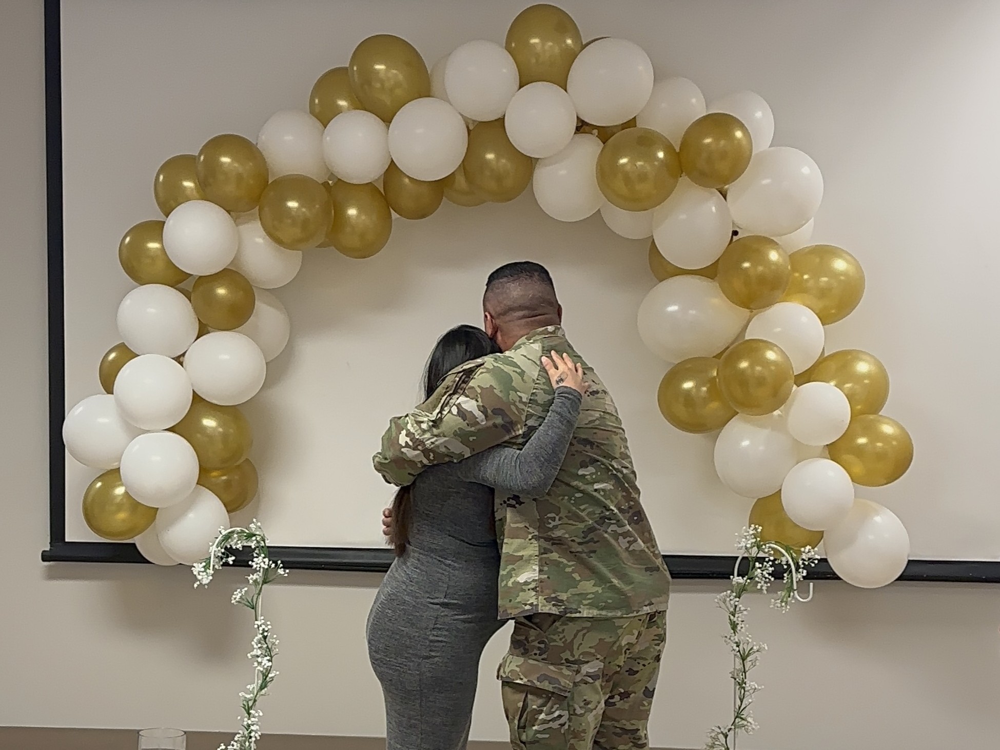 Two people embrace beneath a white and gold balloon arch set up indoors. Decorative floral arrangements sit on stands nearby, creating a wedding-style backdrop in a hospital room setting.