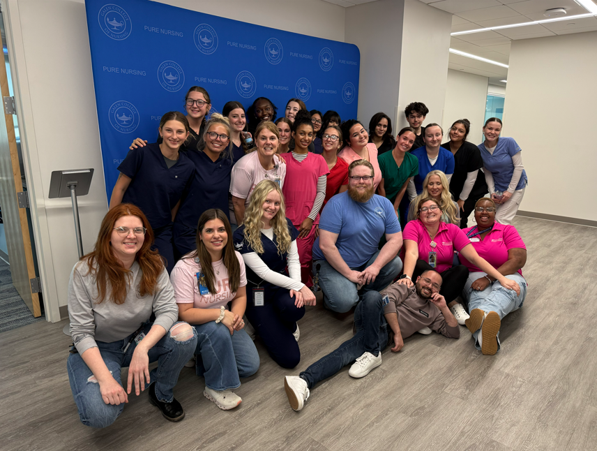 Large group of students and faculty pose together indoors in front of a blue “Pure Nursing” backdrop.