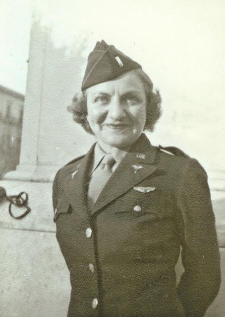 Vintage photograph of a person in a military uniform with insignia and a side cap, standing outdoors near a stone wall or column. The uniform jacket is buttoned, and wings and badges are visible on the chest.