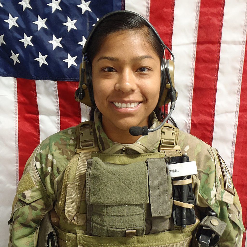 Color photograph of a person wearing a camouflage military uniform and protective vest with attached radio equipment and a headset with microphone. The person stands in front of a large American flag that fills the background.