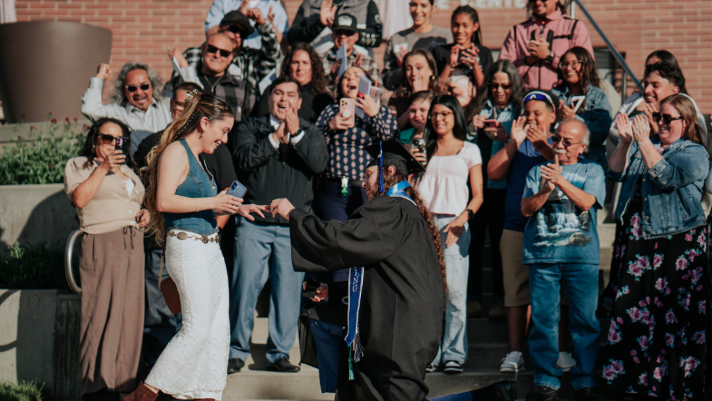 A person in a graduation cap and gown kneels on one knee to place a ring on another person’s hand outdoors, while a large group of people behind them stand on steps clapping and holding phones. A brick building in the background includes partial visible text reading “Central Miller … Center.”
