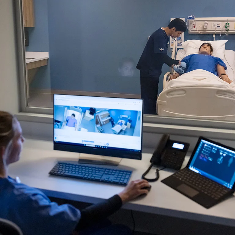 A student in blue scrubs sits at a desk with two computer monitors and a phone, observing a simulation. Through a large window, another student in blue scrubs is seen attending to a medical manikin in a hospital bed, simulating a clinical scenario.