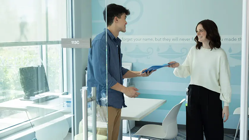 A female admissions counselor meeting with a young man in a sunlit office at Galen, handing him a checklist.