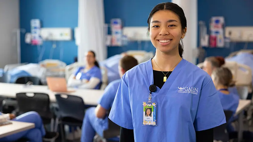 Female nursing student smiling at camera with simulation lab activities happening behind her.