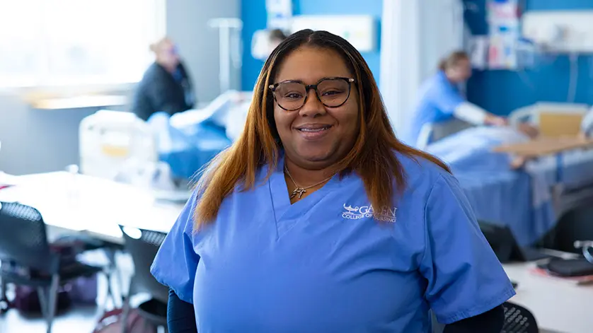 Female nursing student smiling at camera with simulation lab activities happening behind her.
