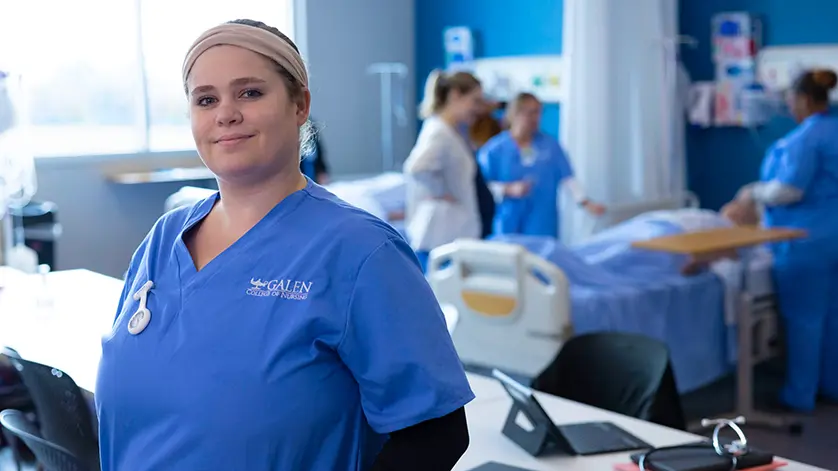 Female nursing student looking at camera with simulation lab activities happening behind her.