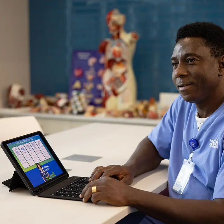 A male Galen student in scrubs looks up from his laptop while studying in a classroom with anatomical models in the background