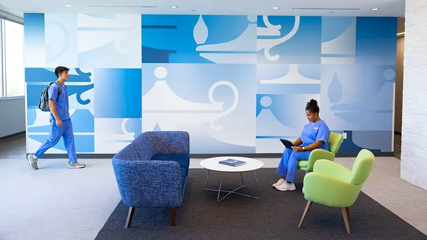Galen student study area, with a male student in scrubs walking along a wall covered in the Galen lamp logo, while a female student also in scrubs sits in a chair and looks at her laptop