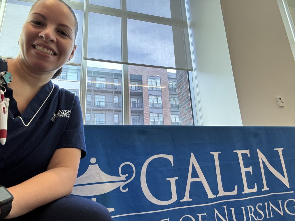 Healthcare worker wearing scrubs and an ID badge stands indoors near large windows, with a blue Galen College banner in the foreground and apartment buildings visible outside.