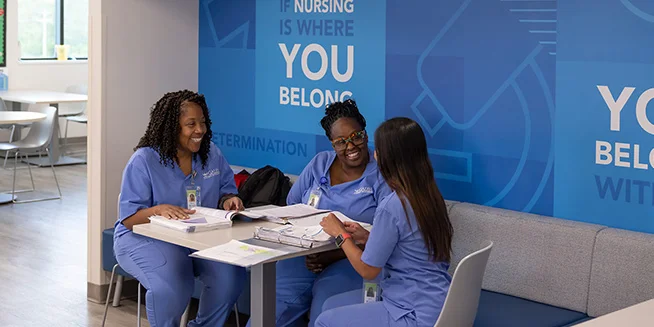 Three smiling female students in blue scrubs are seated at a table, studying together with books and papers spread out. Behind them is a blue wall with inspirational text about nursing.