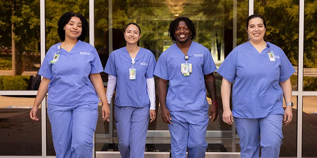 Four medical professionals, two men and two women, all wearing light blue scrubs and identification badges, walk towards the viewer with smiles on their faces. They are in an outdoor foyer with trees visible in the background.