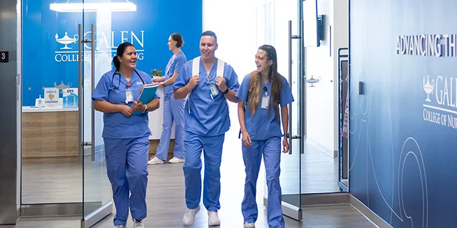 Three nursing students in blue scrubs are walking down a brightly lit hallway in a modern academic building. One woman is smiling and carrying a book, while the other two, a man and a woman, are also smiling and engaged in conversation. The walls are blue with Galen College of Nursing branding.